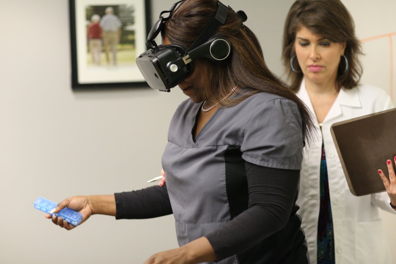 A caregiver wearing the simulator headset reaches out with a pill box while an observer with a clipboard watches